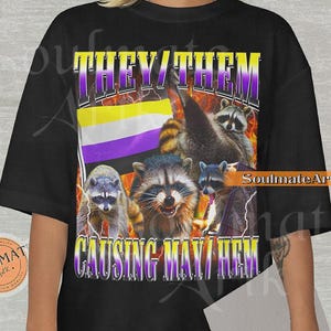 May include: Black t-shirt featuring a graphic of raccoons, the non-binary pride flag, and the text "THEY/THEM CAUSING MAYHEM" in a purple and yellow design. The shirt has a crew neck and short sleeves.