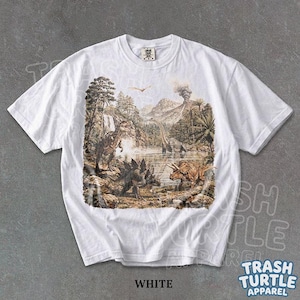 May include: White t-shirt featuring a detailed prehistoric landscape with various dinosaurs, including a Tyrannosaurus Rex, Stegosaurus, and Triceratops. The graphic includes a volcano, trees, and a lake. The text "WHITE" is at the bottom.