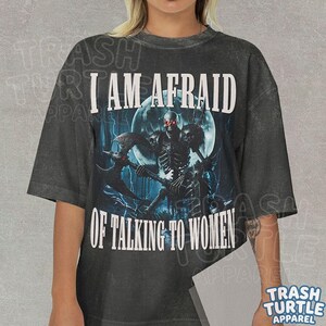 May include: A charcoal gray short-sleeve t-shirt featuring a graphic of a skeleton warrior holding an axe in front of a full moon. The text "I AM AFRAID OF TALKING TO WOMEN" is printed in white capital letters above and below the central illustration.
