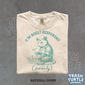 Könnte beinhalten: Beigefarbenes T-Shirt mit einem tealfarbenen Aufdruck eines Opossums mit Hammer und dem Text "I'M BUILT DIFFERENT (poorly)". Das Shirt trägt auch den Text "Trash Turtle Apparel".