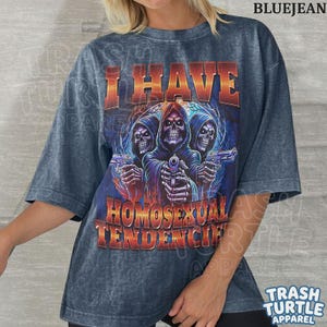 Peut inclure: Un t-shirt gris fonc&eacute; d&eacute;lav&eacute; &agrave; l'acide avec un motif de trois squelettes &agrave; capuche tenant des pistolets. Le texte I HAVE HOMOSEXUAL TENDENCIES est imprim&eacute; dans une police d&eacute;grad&eacute;e orange et jaune au-dessus et en dessous de l'illustration.