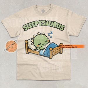 Puede incluir: Camiseta beige con un dinosaurio de dibujos animados durmiendo en una cama. El texto "SLEEPYSAURUS" está encima del dinosaurio. El dinosaurio es verde con una almohada blanca y una manta azul. La camiseta tiene cuello redondo.