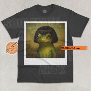 May include: Dark gray t-shirt featuring a framed image of a green frog with a black bob haircut. The frog has large, expressive eyes. The shirt has a round neck and short sleeves. The text "SoulmateArtK" is visible.