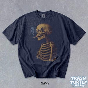 Puede incluir: Camiseta azul marino con una ilustraci&oacute;n detallada de un esqueleto fumando un cigarrillo. El esqueleto se representa de perfil, con detalles anat&oacute;micos. La camiseta tiene el texto "NAVY" en la parte inferior. La marca "TRASH TURTLE APPAREL" tambi&eacute;n es visible.