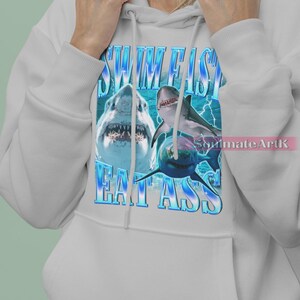 May include: White hoodie featuring a graphic of sharks and the text "SWIM FISH EAT ASS" in blue. The design includes images of sharks with open mouths and a blue background.
