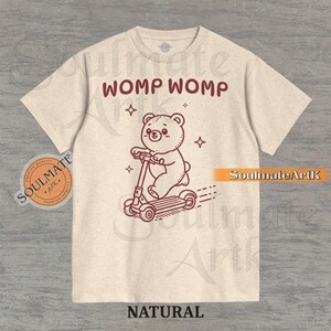 Puede incluir: Camiseta de color natural con una ilustración marrón de un oso en un patinete, con las palabras "WOMP WOMP" arriba y "NATURAL" abajo. La camiseta también tiene un logotipo circular en el lado izquierdo.