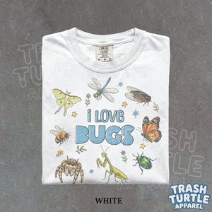 Peut inclure: Un t-shirt blanc avec une impression graphique colorée représentant divers insectes, dont un papillon de nuit, une libellule, une cigale, une abeille, une araignée, une mante et un scarabée. Le texte "i LOVE BUGS" est imprimé en grandes lettres bleues.