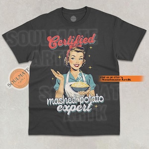 Può includere: T-shirt grigio scuro con un'illustrazione retrò di una donna che tiene in mano purè di patate. Il testo recita "Certified mashed potato expert". Il design include una fascia rossa e un grembiule.
