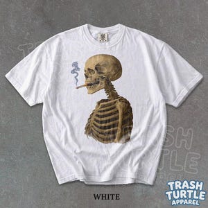 Puede incluir: Camiseta blanca con una ilustraci&oacute;n detallada de un esqueleto fumando un cigarrillo. El esqueleto est&aacute; representado en tonos marrones y beige, con humo azul. La camiseta tiene cuello redondo y mangas cortas.