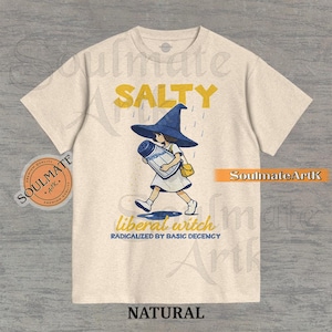 May include: Natural-colored t-shirt featuring a cartoon witch carrying a salt shaker under a blue hat. The text "SALTY" is above the witch, and "liberal witch RADICALIZED BY BASIC DECENCY" is below. The shirt has a round neck.