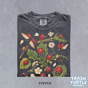 Op de afbeelding: Een antracietgrijs t-shirt met korte mouwen en een grafische print van rode lieveheersbeestjes, groene eikenbladeren en witte bloemen. Het shirt heeft een ronde hals en een label met de tekst Comfort Colors. Het ontwerp staat op de voorzijde.