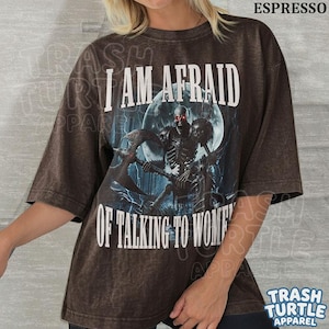 May include: A dark brown, short-sleeved t-shirt featuring a graphic of a skeleton holding an axe in front of a full moon. The text reads I AM AFRAID OF TALKING TO WOMEN in white capital letters. The shirt has a distressed, vintage-style wash.