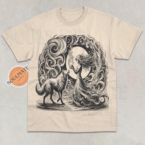 Puede incluir: Una camiseta beige con un estampado gráfico en blanco y negro de una mujer con el pelo largo y un lobo. La mujer sostiene una llama en su mano y el lobo está de pie a su lado. El gráfico está rodeado por un diseño en espiral.