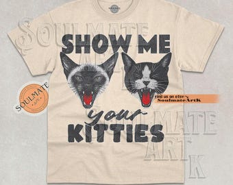 May include: Cream-colored t-shirt featuring two cat faces with open mouths and the text "SHOW ME your KITTIES" in black. The cat faces are black and white with red mouths. The shirt has a round neckline.