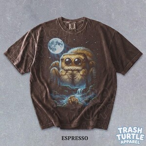 May include: A brown short-sleeve t-shirt featuring a graphic of a large, fuzzy jumping spider with big eyes under a full moon. Below the spider, a smaller spider sits near a waterfall in a forest setting. The shirt has a tag labeled Trash Turtle.