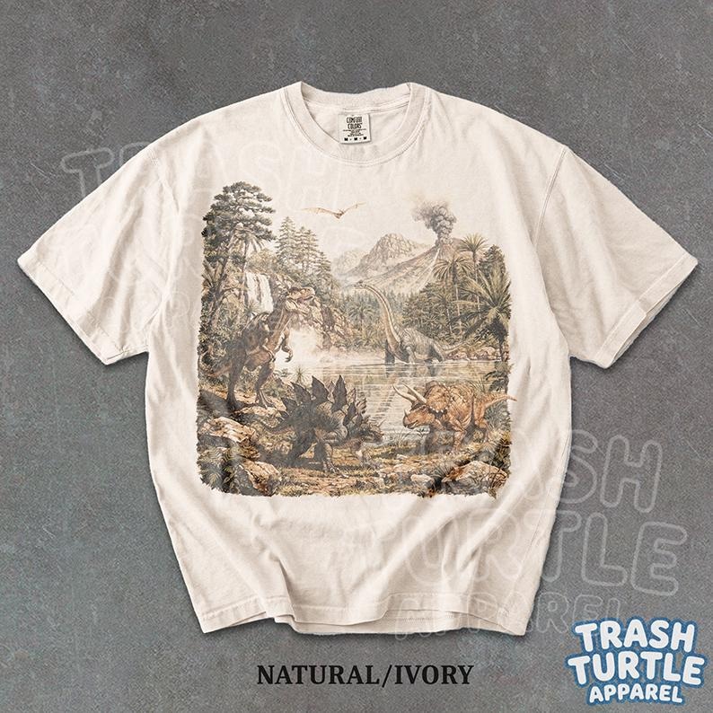 May include: Ivory-colored t-shirt featuring a detailed illustration of dinosaurs in a prehistoric landscape. The design includes various dinosaur species, trees, mountains, and a volcano. The text "NATURAL/IVORY" is printed at the bottom.