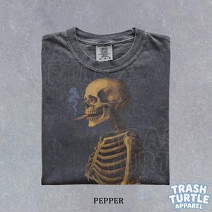 Puede incluir: Camiseta gris oscuro con una ilustraci&oacute;n detallada de un esqueleto fumando un cigarrillo. El esqueleto est&aacute; representado en tonos marrones y beige, con humo azul. La camiseta tiene el texto "PEPPER" y "TRASH TURTLE APPAREL".