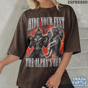 May include: Espresso-colored t-shirt featuring a werewolf graphic with the text "HIDE YOUR FEET" and "THE ALPHA'S HERE". The design includes a full moon and flames. The shirt is from Trash Turtle Apparel.