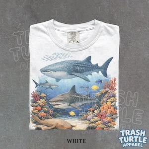 Puede incluir: Camiseta blanca con un diseño de vida marina, que incluye tiburones, corales y peces. El gráfico presenta un tiburón ballena, un tiburón tigre y otras criaturas marinas. La camiseta tiene cuello redondo y la palabra "WHITE" está impresa en la parte inferior.