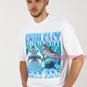 May include: White t-shirt featuring a graphic of three sharks with the text "SWIM FAST EAT ASS" in blue. The design includes lightning bolts and a gradient effect, creating a bold and eye-catching design. The shirt is a casual wear item.
