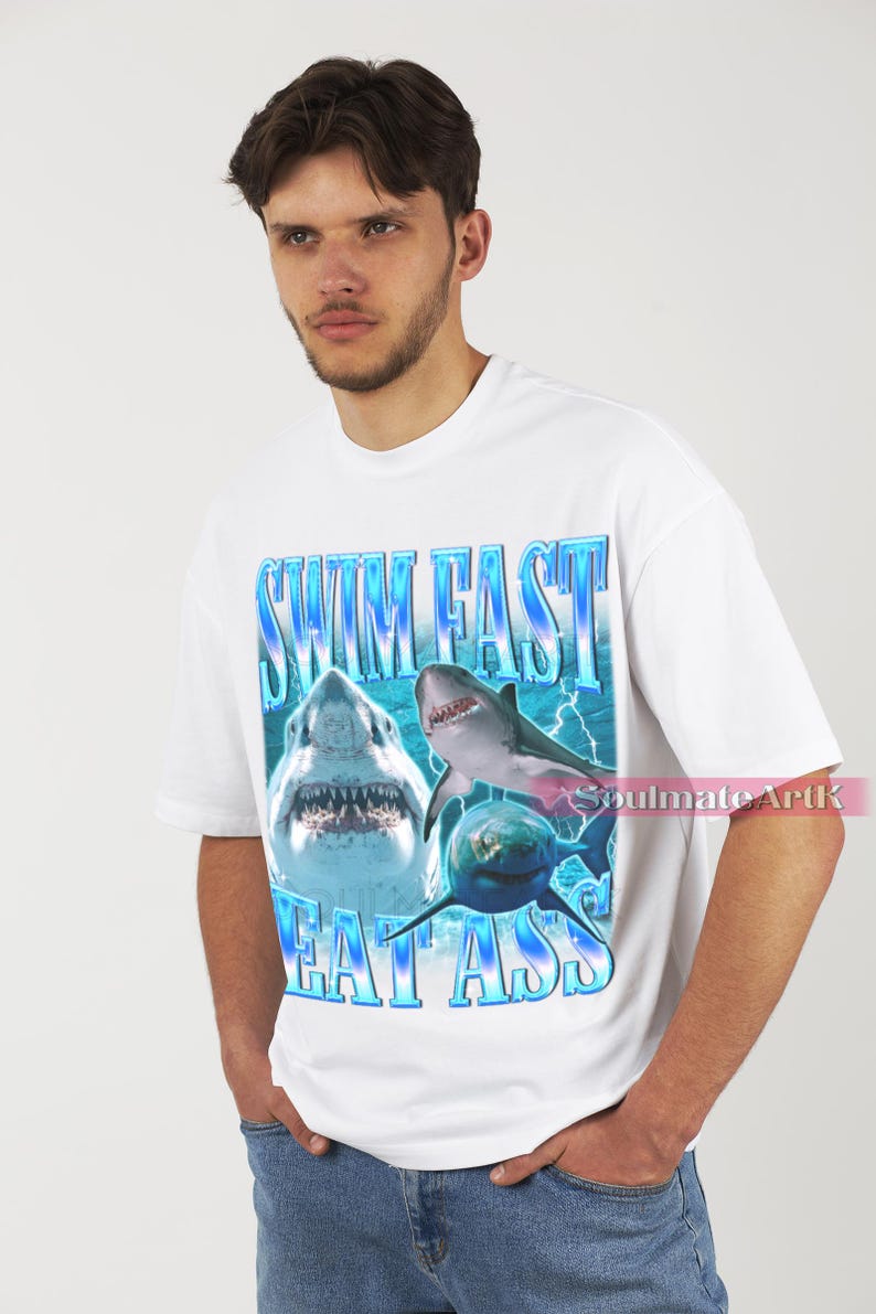 May include: White t-shirt featuring a graphic of three sharks with the text "SWIM FAST EAT ASS" in blue. The design includes lightning bolts and a gradient effect, creating a bold and eye-catching design. The shirt is a casual wear item.