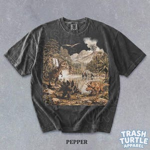 May include: Pepper-colored t-shirt featuring a detailed prehistoric landscape with various dinosaurs, trees, and a volcano. The shirt has a vintage wash and the text "TRASH TURTLE APPAREL" is visible.