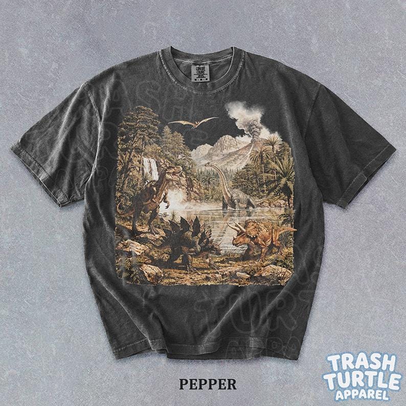 May include: Pepper-colored t-shirt featuring a detailed prehistoric landscape with various dinosaurs, trees, and a volcano. The shirt has a vintage wash and the text "TRASH TURTLE APPAREL" is visible.