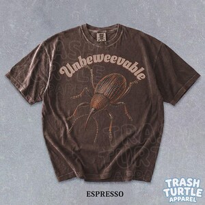 Puede incluir: Una camiseta de manga corta de color marr&oacute;n oscuro con un gr&aacute;fico de un gorgojo en el centro. El texto "Unbeweevable" est&aacute; impreso en una fuente clara y curva sobre el insecto. La camiseta tiene cuello redondo y un acabado desgastado.
