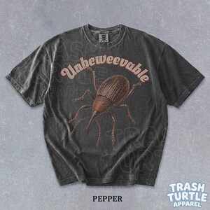 Puede incluir: Una camiseta de manga corta gris carb&oacute;n con una ilustraci&oacute;n marr&oacute;n de un insecto gorgojo centrada en el pecho. Sobre el insecto, la palabra Unbeweevable est&aacute; impresa en una fuente curva de color claro. La camiseta tiene cuello redondo.