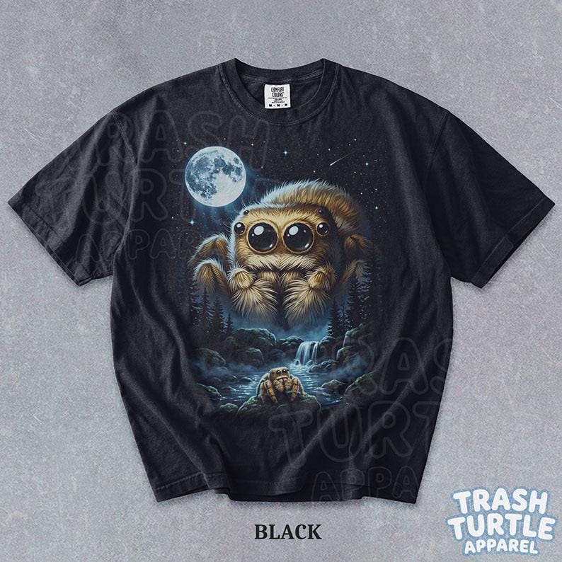 May include: A black short-sleeve t-shirt featuring a graphic print of a large, fluffy, brown jumping spider with oversized eyes floating above a dark forest landscape with a waterfall and a full moon in the starry night sky above.