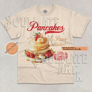 May include: A cream-coloured t-shirt with the word "Pancakes" in red script above a stack of pancakes topped with strawberries and banana slices. The shirt also features the text "& Panic Attacks!" and "100% Organic Anxiety."