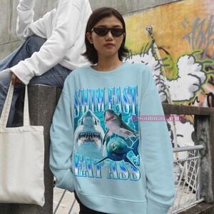 May include: Light blue sweatshirt with the text "SWIM FAST EAT ASS" and a graphic of three sharks. The sweatshirt is a crew neck style and has a vintage aesthetic. The text and graphic are in shades of blue and white.