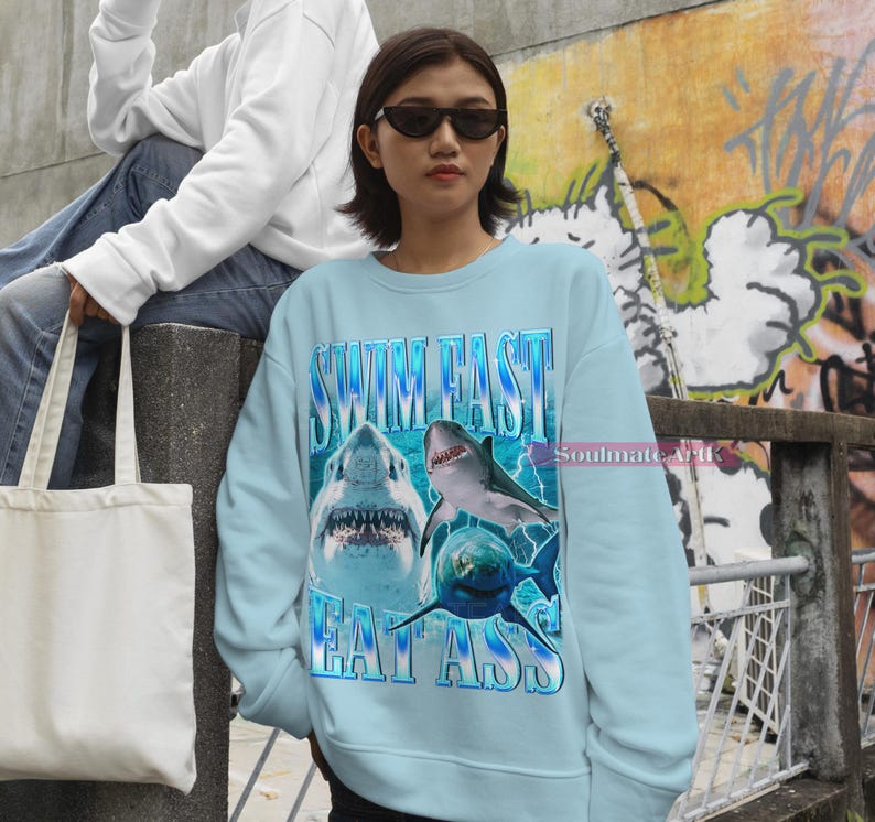 May include: Light blue sweatshirt with the text "SWIM FAST EAT ASS" and a graphic of three sharks. The sweatshirt is a crew neck style and has a vintage aesthetic. The text and graphic are in shades of blue and white.