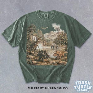 May include: Military green t-shirt with a detailed dinosaur landscape graphic. The design features various dinosaurs, trees, a volcano, and a lake. The shirt has a crew neck and short sleeves. The text "MILITARY GREEN/MOSS" is printed at the bottom.