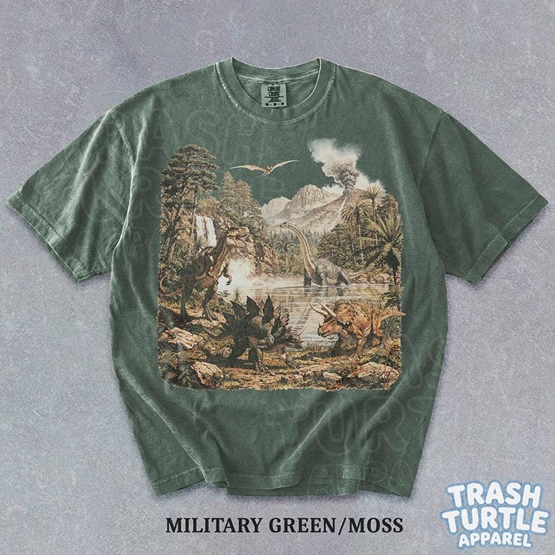 May include: Military green t-shirt with a detailed dinosaur landscape graphic. The design features various dinosaurs, trees, a volcano, and a lake. The shirt has a crew neck and short sleeves. The text "MILITARY GREEN/MOSS" is printed at the bottom.