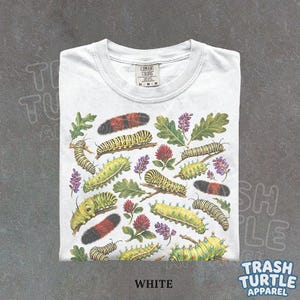 May include: A white t-shirt featuring a detailed botanical and entomological print of various caterpillars, green leaves, and purple and red clover flowers. The shirt has a Comfort Colors brand label at the inner neckline and is size M.