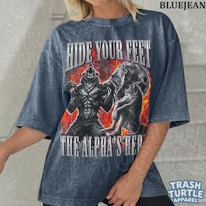 May include: Blue jean-colored t-shirt with a graphic of a werewolf transforming, with the text "HIDE YOUR FEET" and "THE ALPHA'S HERE". The design features a full moon and a fiery background. The shirt is from Trash Turtle Apparel.