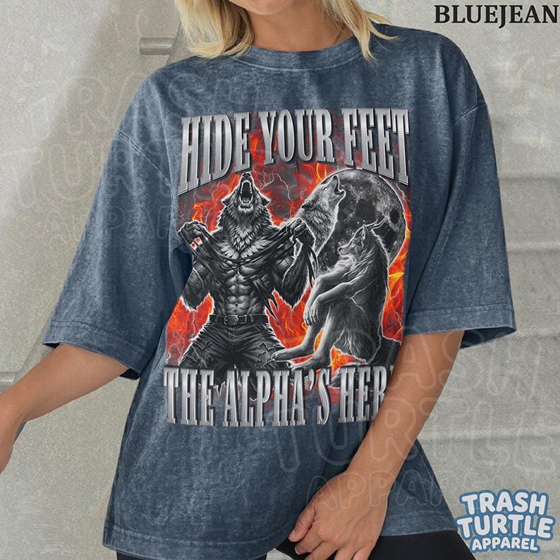 May include: Blue jean-colored t-shirt with a graphic of a werewolf transforming, with the text "HIDE YOUR FEET" and "THE ALPHA'S HERE". The design features a full moon and a fiery background. The shirt is from Trash Turtle Apparel.