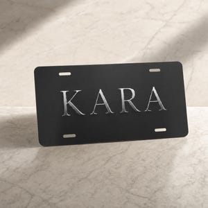 May include: A rectangular black license plate featuring the name KARA in large, metallic silver, serif-style capital letters centered on the front. The plate includes four horizontal mounting slots, two at the top and two at the bottom.