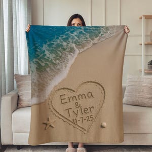 May include: A personalized beach-themed blanket featuring a heart drawn in sand with the names "Emma & Tyler" and the date "11-7-25". The top of the blanket shows a blue ocean wave, and a starfish and seashell are also visible.