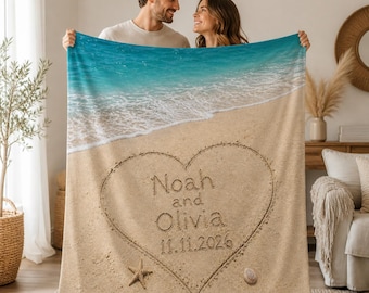 May include: A personalized beach-themed blanket featuring a heart drawn in sand with the names "Noah and Olivia" and the date "11.11.2026". The blanket depicts a turquoise ocean and sandy beach scene.