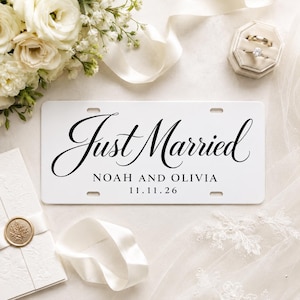 Può includere: Targa bianca con le parole "Just Married" in elegante scrittura nera. Sotto, i nomi "Noah e Olivia" e la data "11.11.26" sono stampati. Circondata da elementi a tema matrimonio.