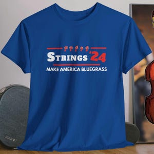 May include: Blue t-shirt with the text "STRINGS '24" and "MAKE AMERICA BLUEGRASS" in red and white. The shirt features a row of red mushrooms above the text.