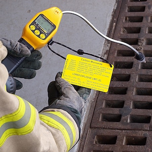 May include: A yellow carbon monoxide detector with a digital display reading 21.0 ppm. The detector is connected to a long, flexible probe. A yellow card with the text "Carbon Monoxide" and safety information is visible in the foreground.