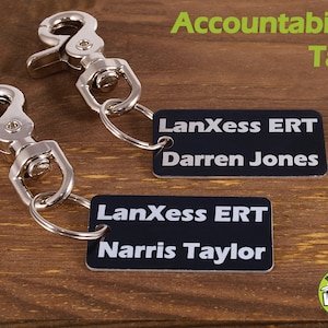 May include: Two black rectangular accountability tags with silver key rings and clips. The tags have white text that reads "LanXess ERT" and "Darren Jones" on one tag and "LanXess ERT" and "Narris Taylor" on the other tag.