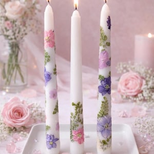 May include: Three lit white pillar candles decorated with pressed flowers in pink, purple, and green. The candles are on a white rectangular tray, with a soft pink background and scattered rose petals. The candles are a decorative home accent.