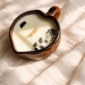 Puede incluir: Una vela en forma de corazón en un recipiente de cerámica con asa. La vela está rellena de cera cremosa y decorada con una piedra negra, un trozo de madera y flores secas. El recipiente tiene rayas marrones y naranjas.