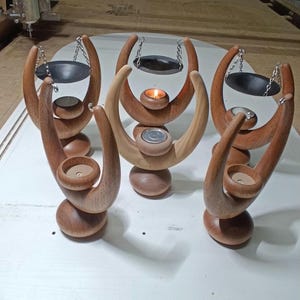 Op de afbeelding: Set van vijf houten kaarsenstandaards met een uniek ontwerp. Elke standaard heeft een gebogen, halvemaanvormige bovenkant met een klein metalen schoteltje dat aan kettingen hangt. De schoteltjes zijn ontworpen om essentiële oliën of wax melts te bevatten. De standaards zijn gemaakt van natuurlijk hout en hebben een warme, rustieke afwerking.