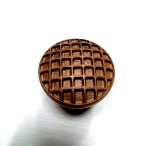 May include: A round, bronze-colored cabinet knob with a waffle-like grid pattern. The knob is made of metal and has a slightly domed shape. The background is white.