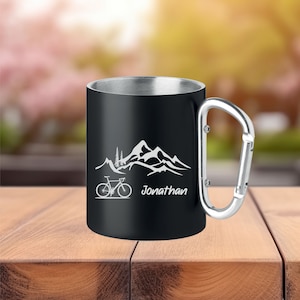 Könnte beinhalten: Schwarze Reise-Thermoskanne aus Edelstahl mit Karabinerhaken. Die Thermoskanne hat ein weißes Design mit einer Bergkette und einem Fahrrad davor. Der Text "Jonathan" ist unter dem Design graviert.
